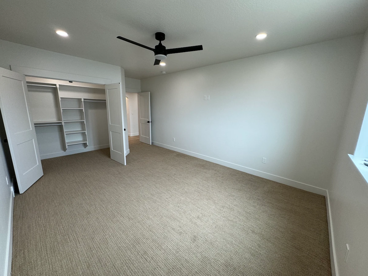 Empty bedroom with beige carpet, white walls, an open closet with shelves, professional ceiling fan installation, recessed lighting, and a window on the right.