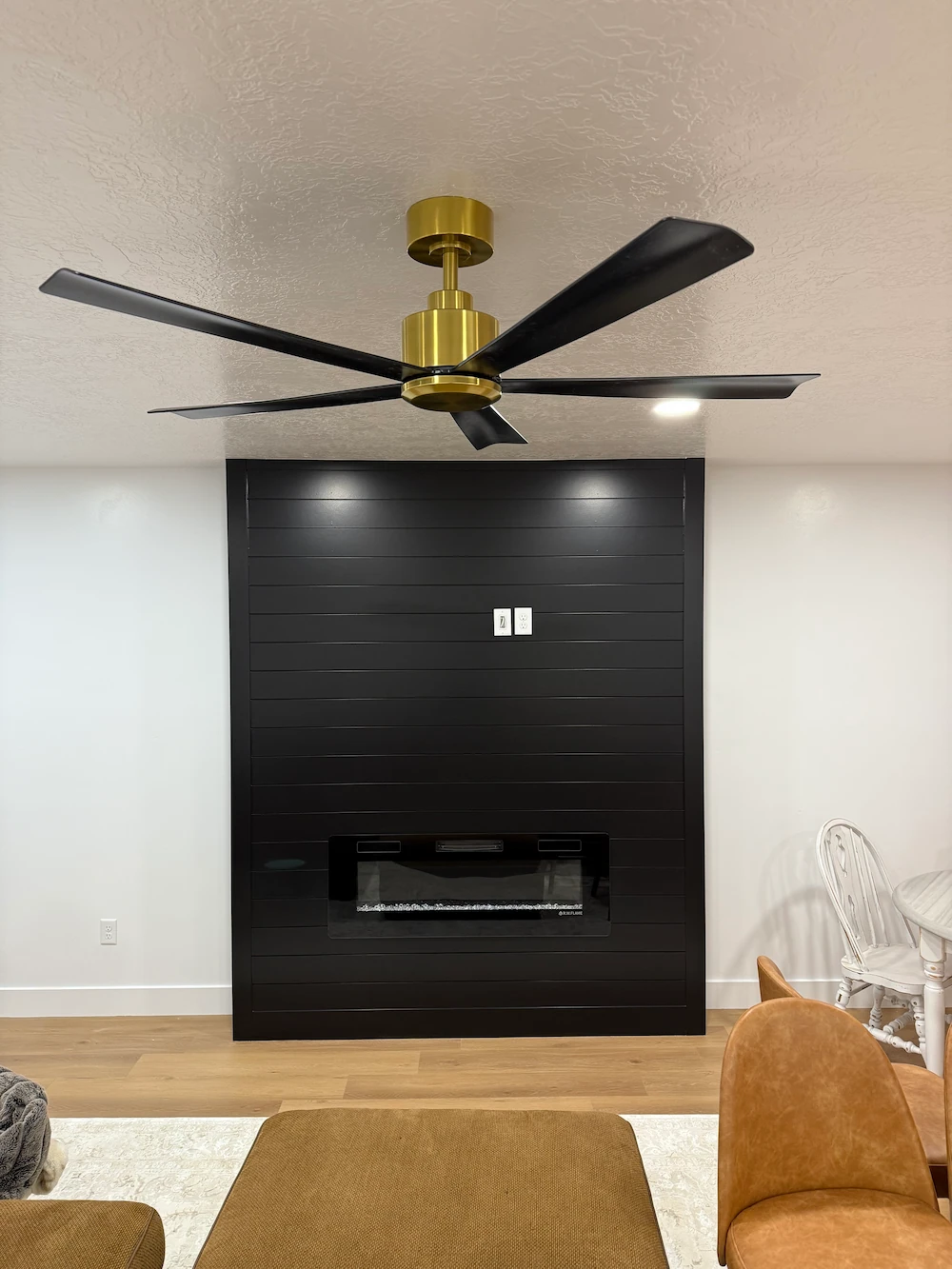 Modern living room with a black accent wall featuring an electric fireplace, professionally done ceiling fan installation in gold and black, wood flooring, and tan furniture.