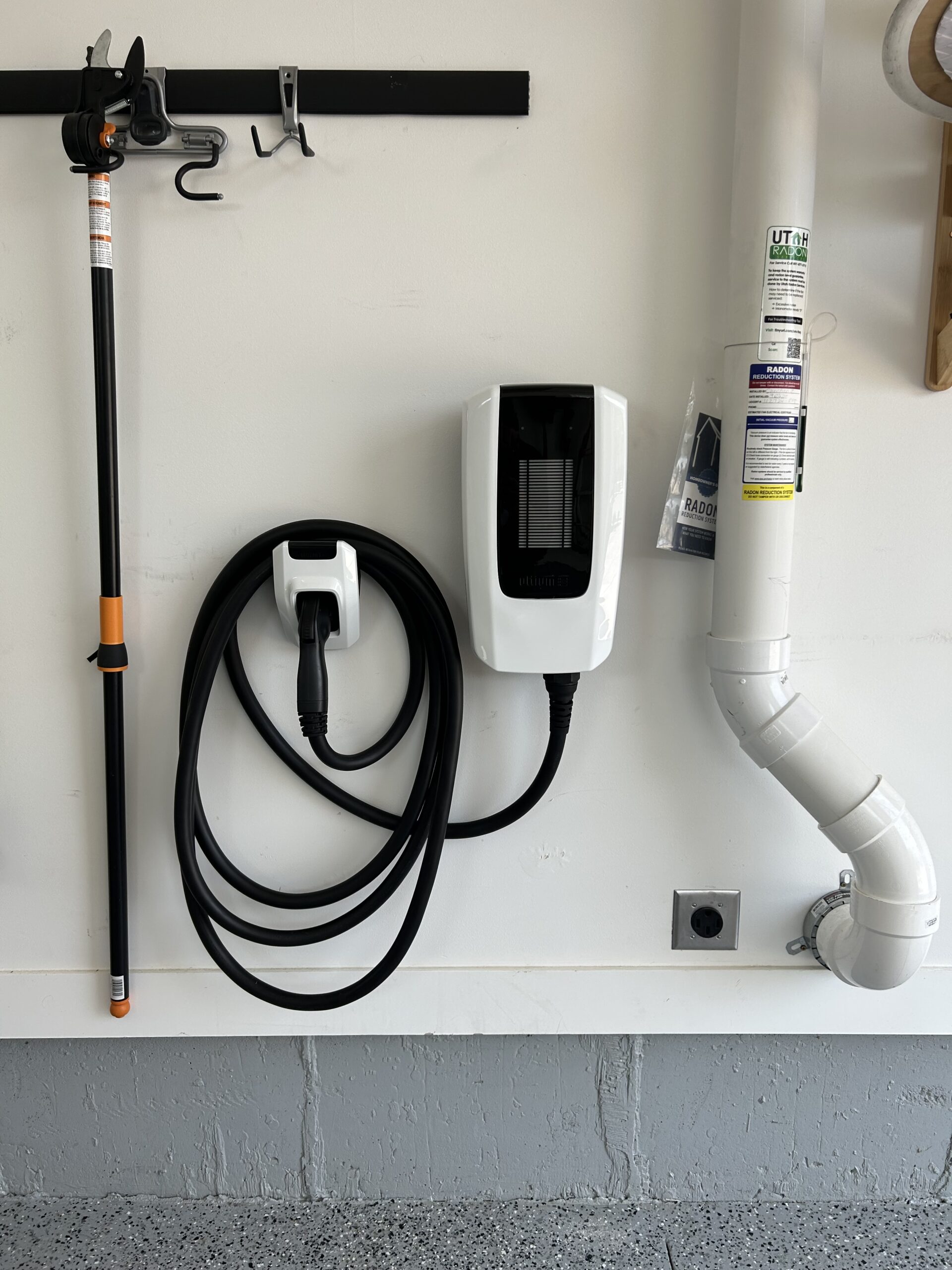 A wall-mounted electric vehicle charging station with a coiled charging cable is installed next to a white pipe in a garage.