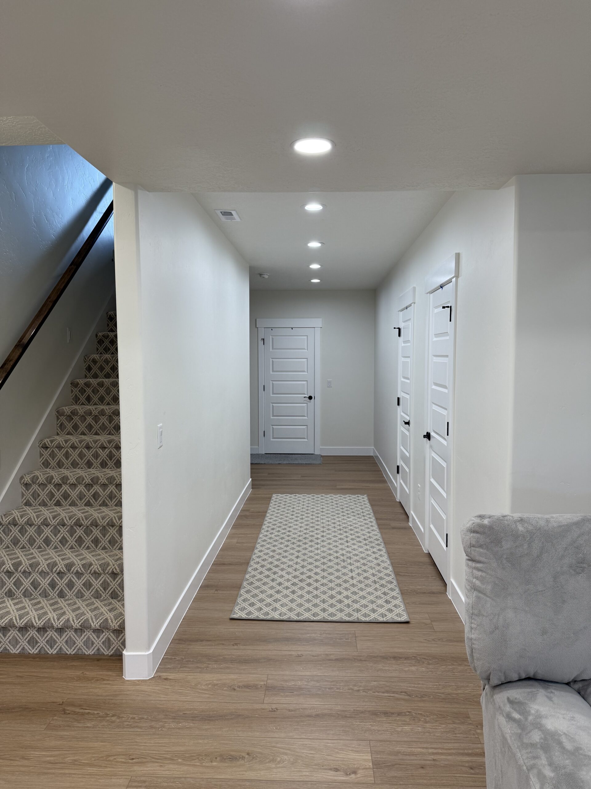 A hallway with wood floors, white walls, three white doors, a patterned carpet runner, and carpeted stairs on the left. Part of a gray sofa is visible in the foreground.