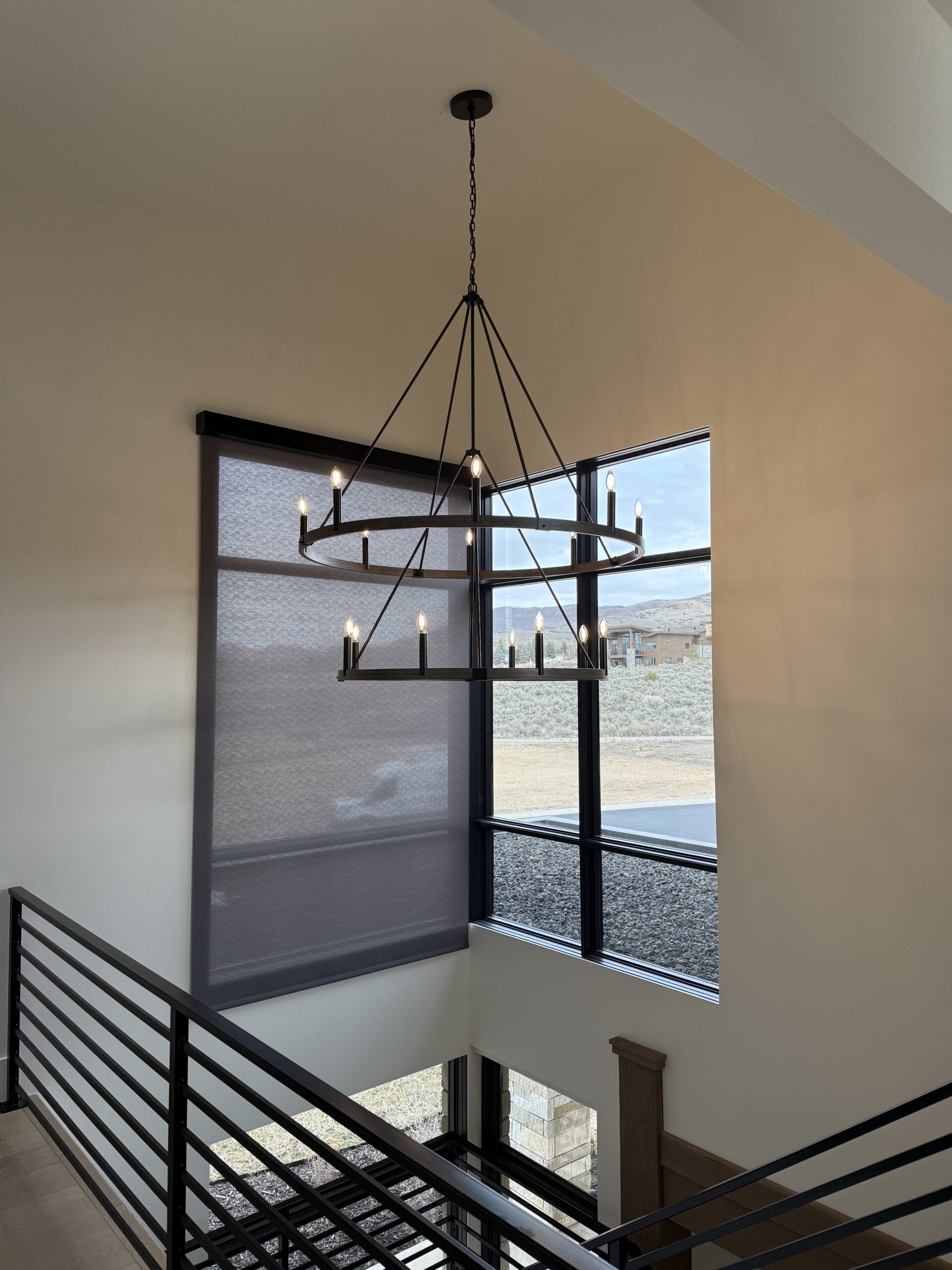 Large black metal chandelier hangs in a modern home with tall windows, letting in natural light and offering a view of an outdoor landscape.