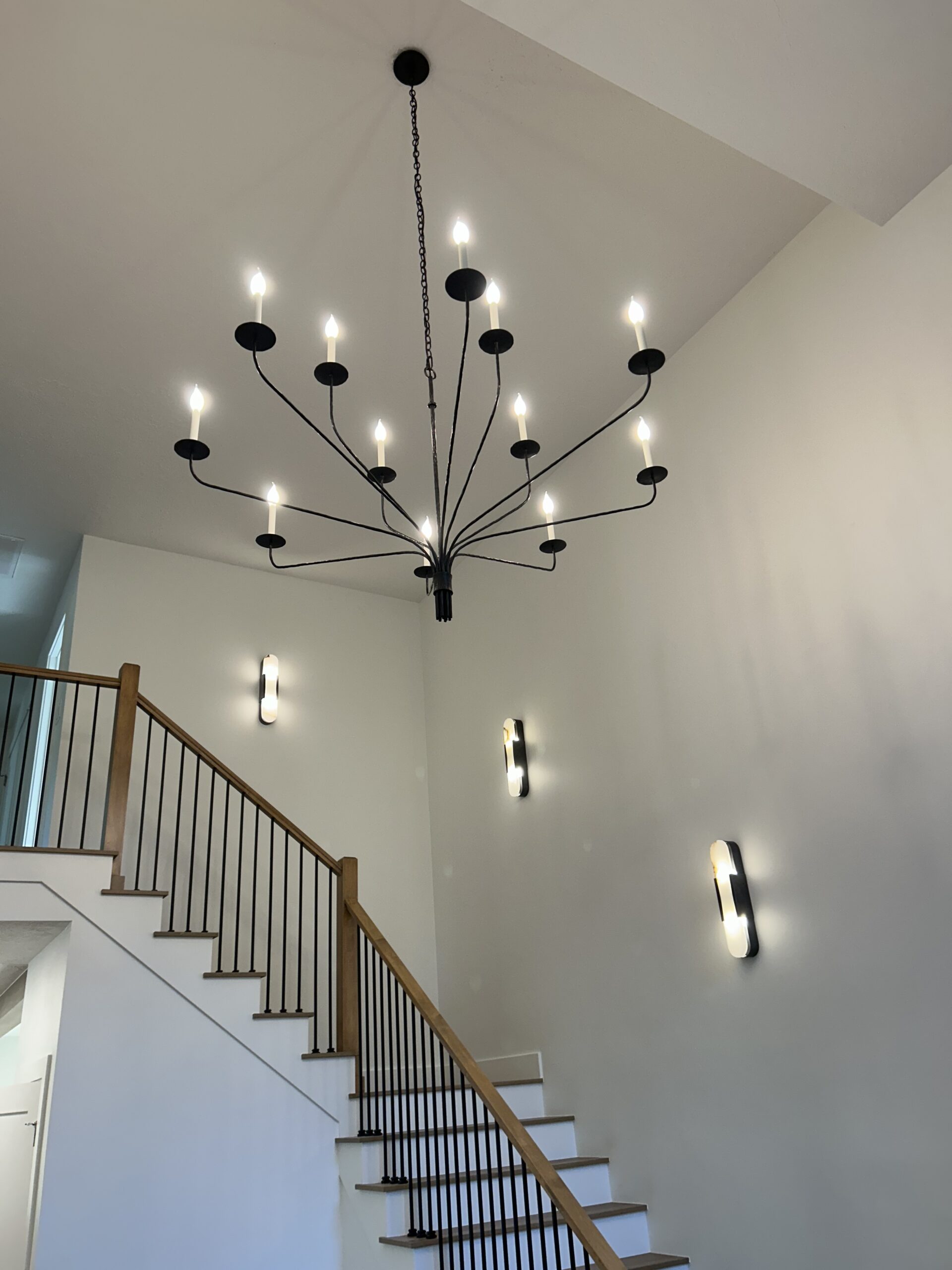 Modern stairwell with a large, black, multi-arm chandelier and three wall-mounted lights; white walls and wooden handrail visible.