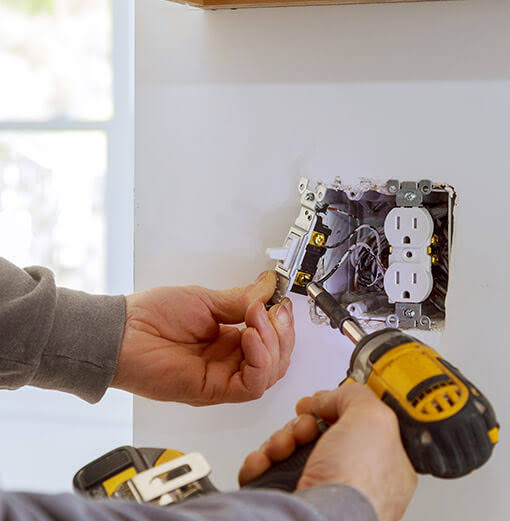 A person uses a power drill to install or remove a light switch next to an electrical outlet on a white wall, possibly preparing for ceiling fan installation.