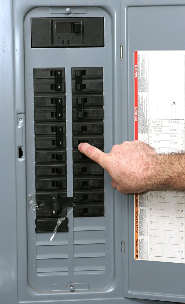 A hand is turning off a circuit breaker inside an electrical panel with labeled switches and a list attached to the door.