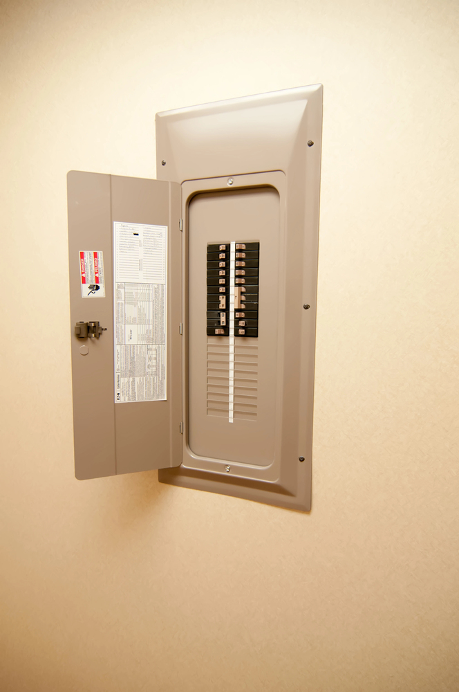 An open electrical breaker panel is mounted on a light-colored wall, showing switches and a label with instructions on the inside of the door.