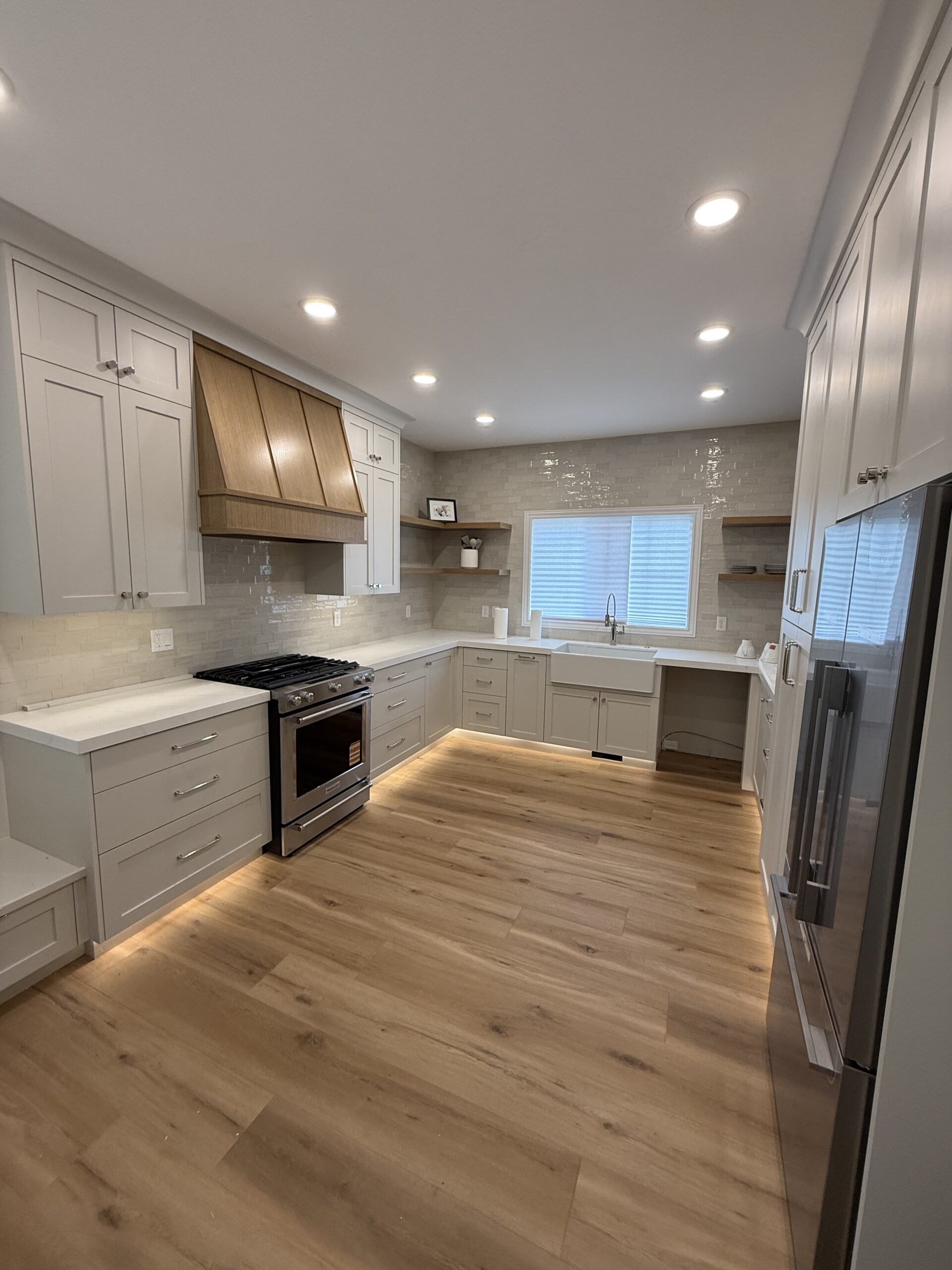 Modern kitchen with light wood flooring, white cabinets, a farmhouse sink, stainless steel appliances, and a wood-accented range hood under recessed lighting.